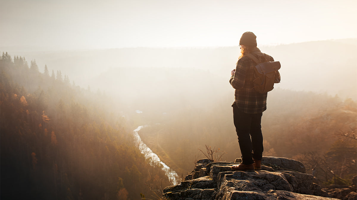 A man standing on the edge of a rocky cliff, looking at the view of a river and forest.