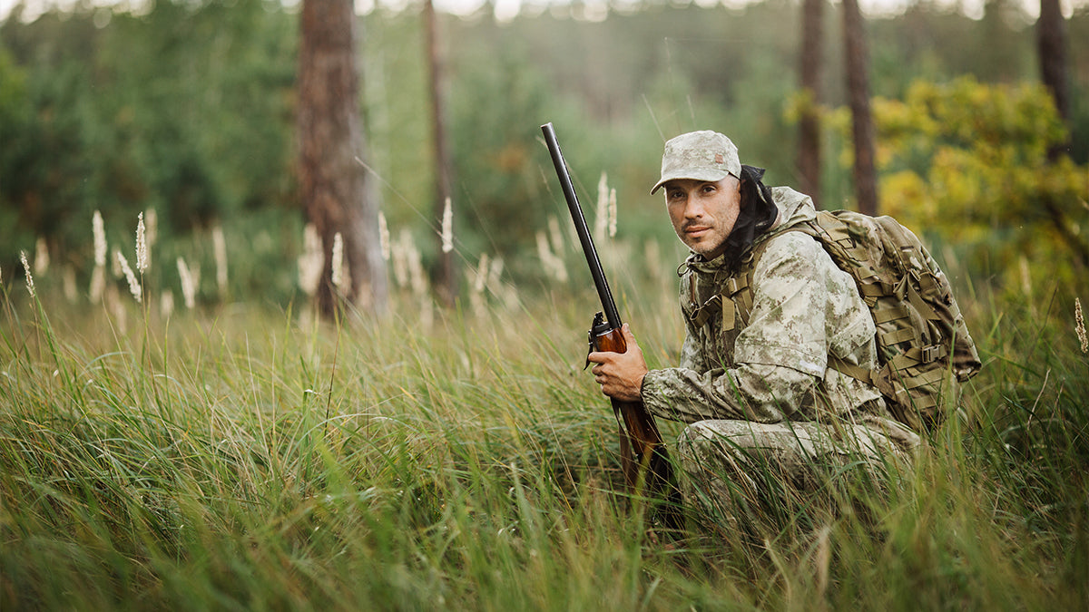 A man dressed in light green camouflage crouching in a green meadow, gun in hand.