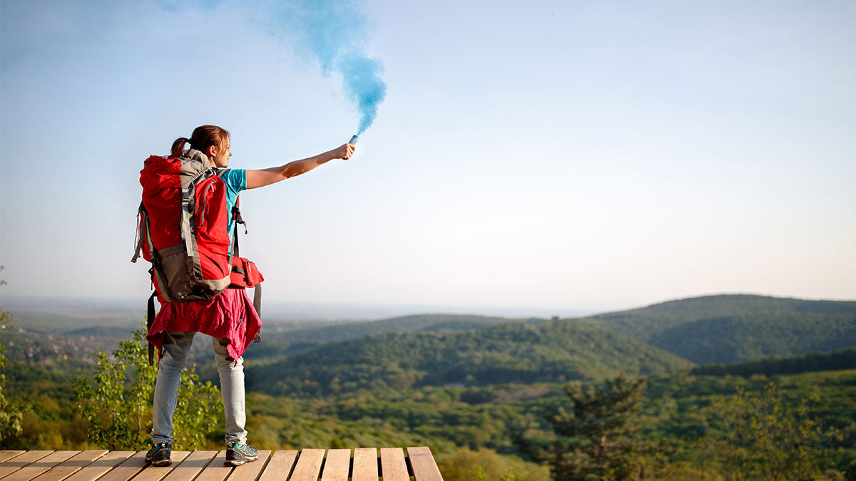 Woman in outdoor gear along the mountainside waving a blue smoke signal.