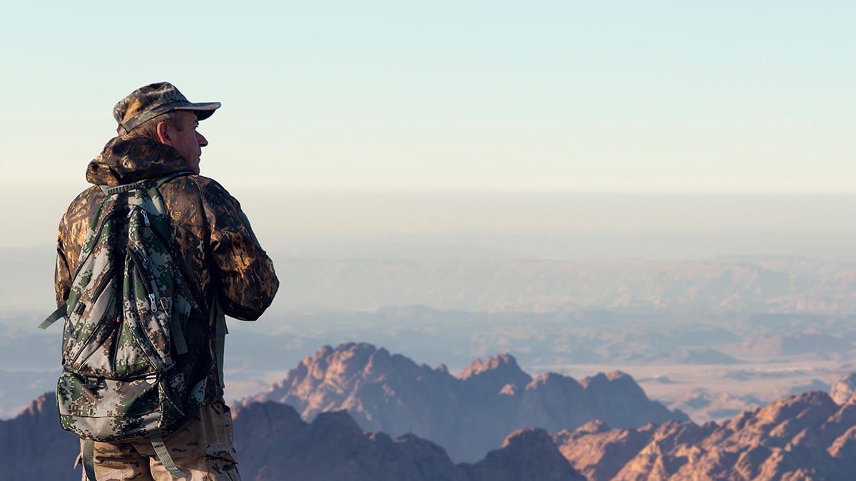 A man wearing camouflage clothing and a camouflage backpack, looking out over the mountains.