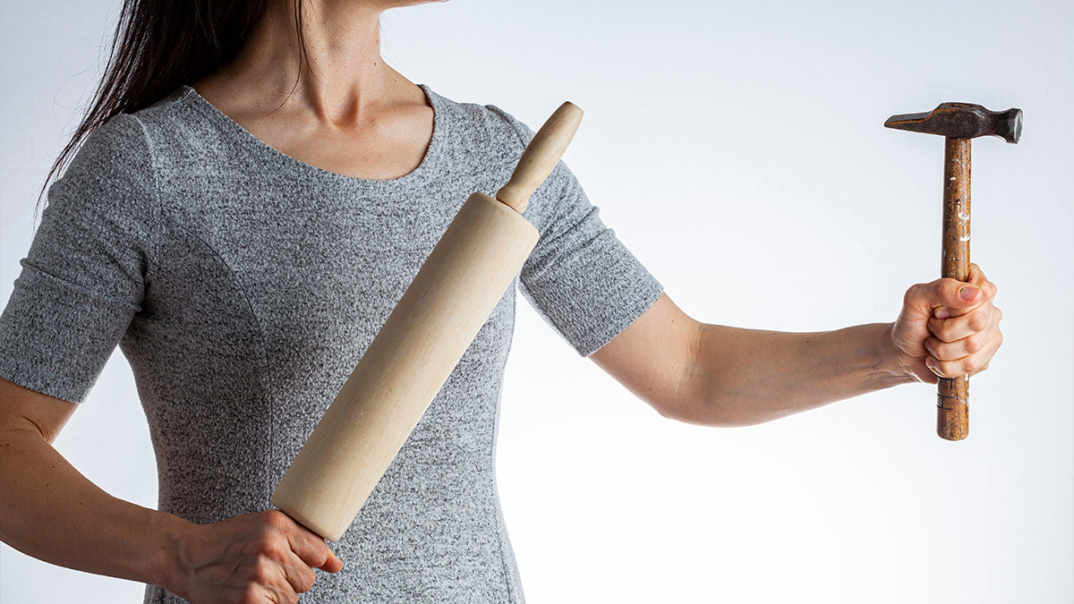 A woman in a gray shirt holding a rolling pin in one hand and a hammer in the other.